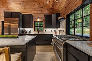 A modern kitchen with dark cabinetry, stainless steel appliances, and a large window offering a view of the forest. - Hemlock Social: Creekside Paradise • Pool • Sauna • HotTub • FirePit • Fun Games (Blue Ridge)