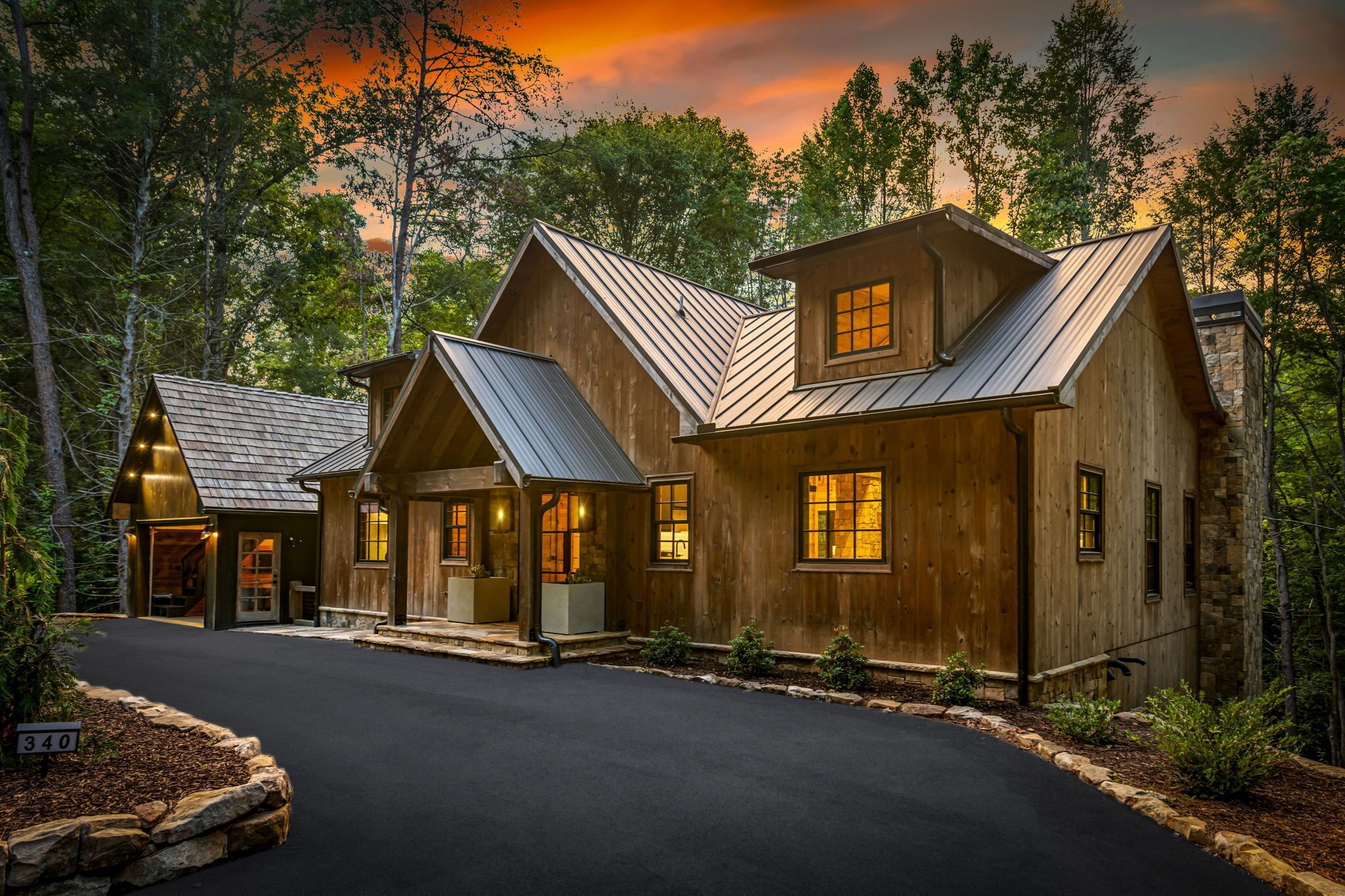 A log cabin with a metal roof, surrounded by trees, and a paved driveway.
