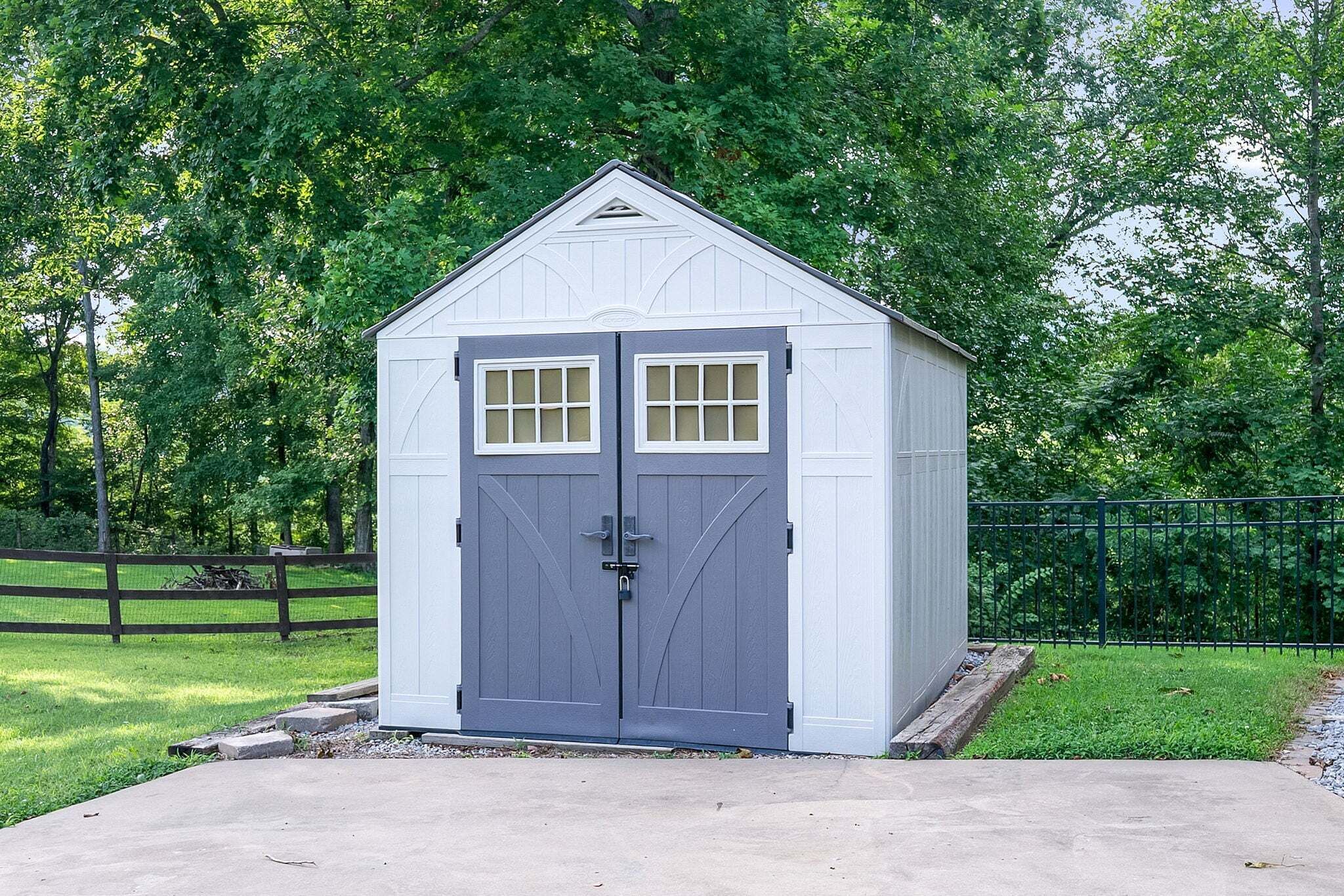A small, single-story shed with a grey door and windows, surrounded by a fence and trees.