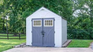 A small, single-story shed with a grey door and windows, surrounded by a fence and trees.