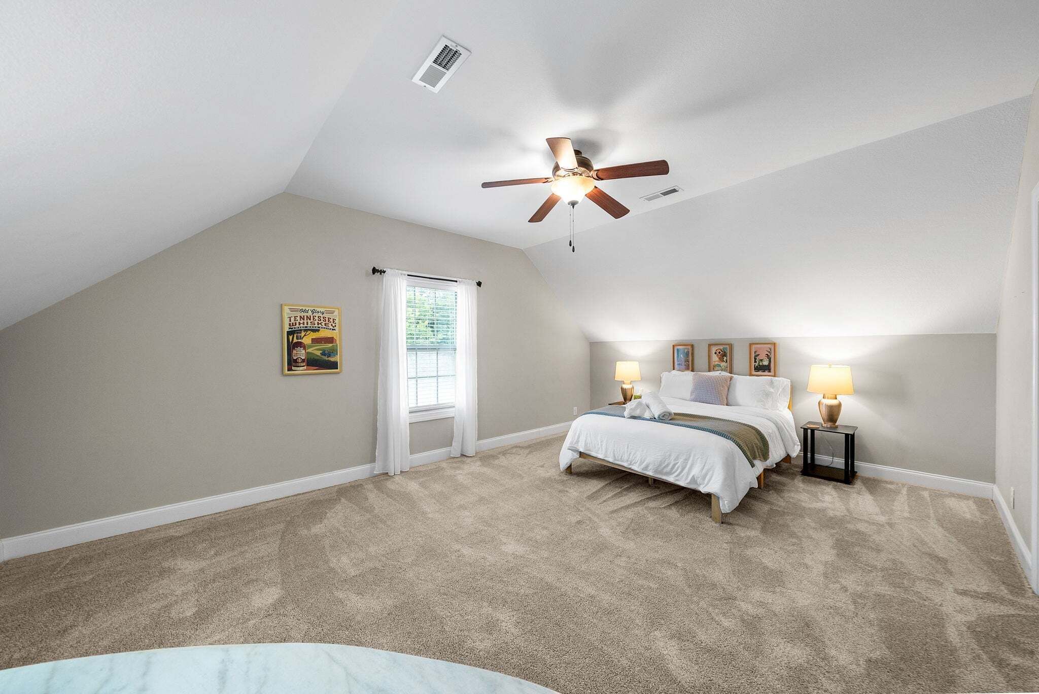 A bedroom with a sloped ceiling, a bed, two bedside tables with lamps, a ceiling fan, and a window with blinds.