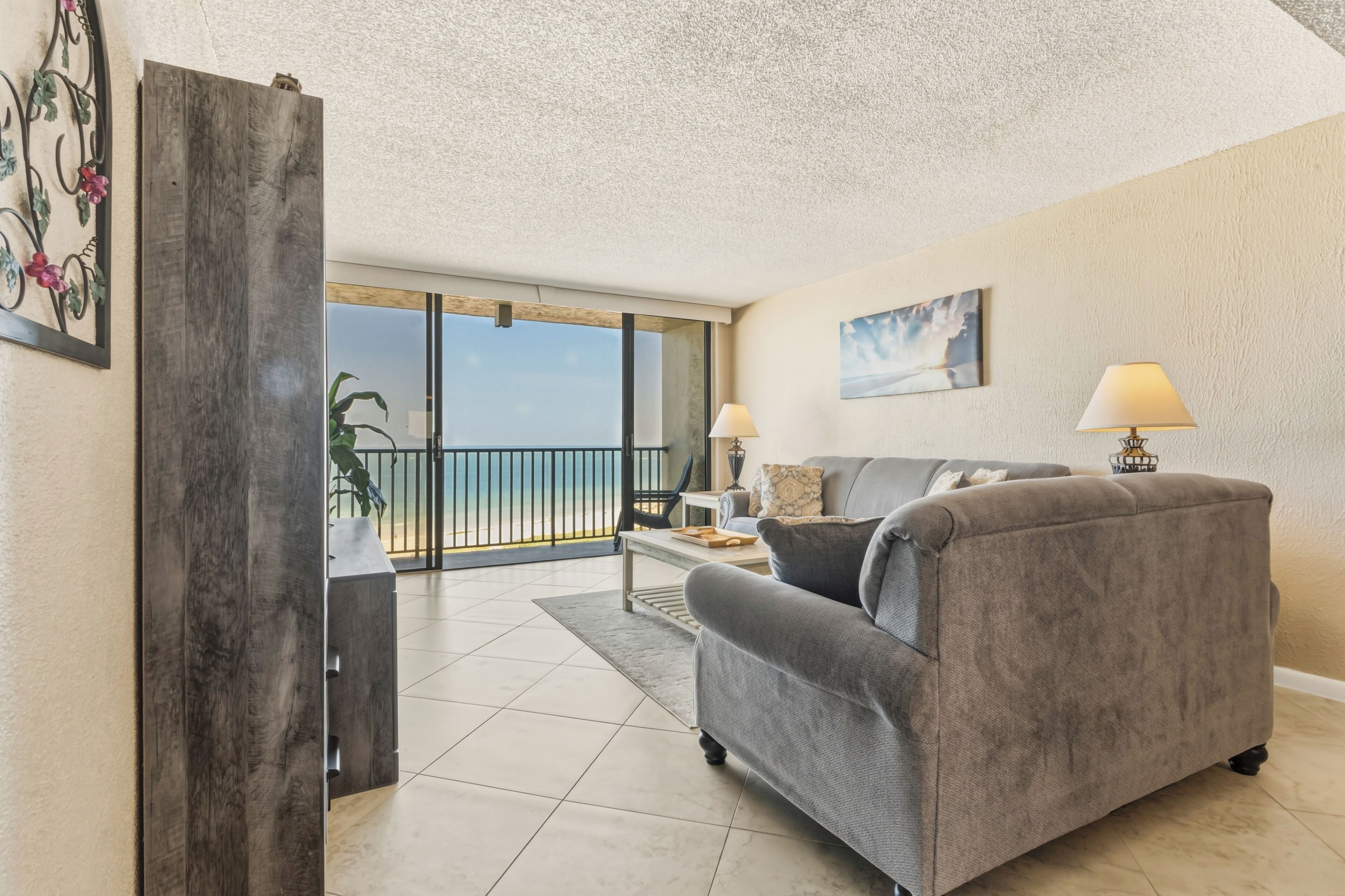 A living room with a grey sofa, two lamps, a painting, and a view of the beach.