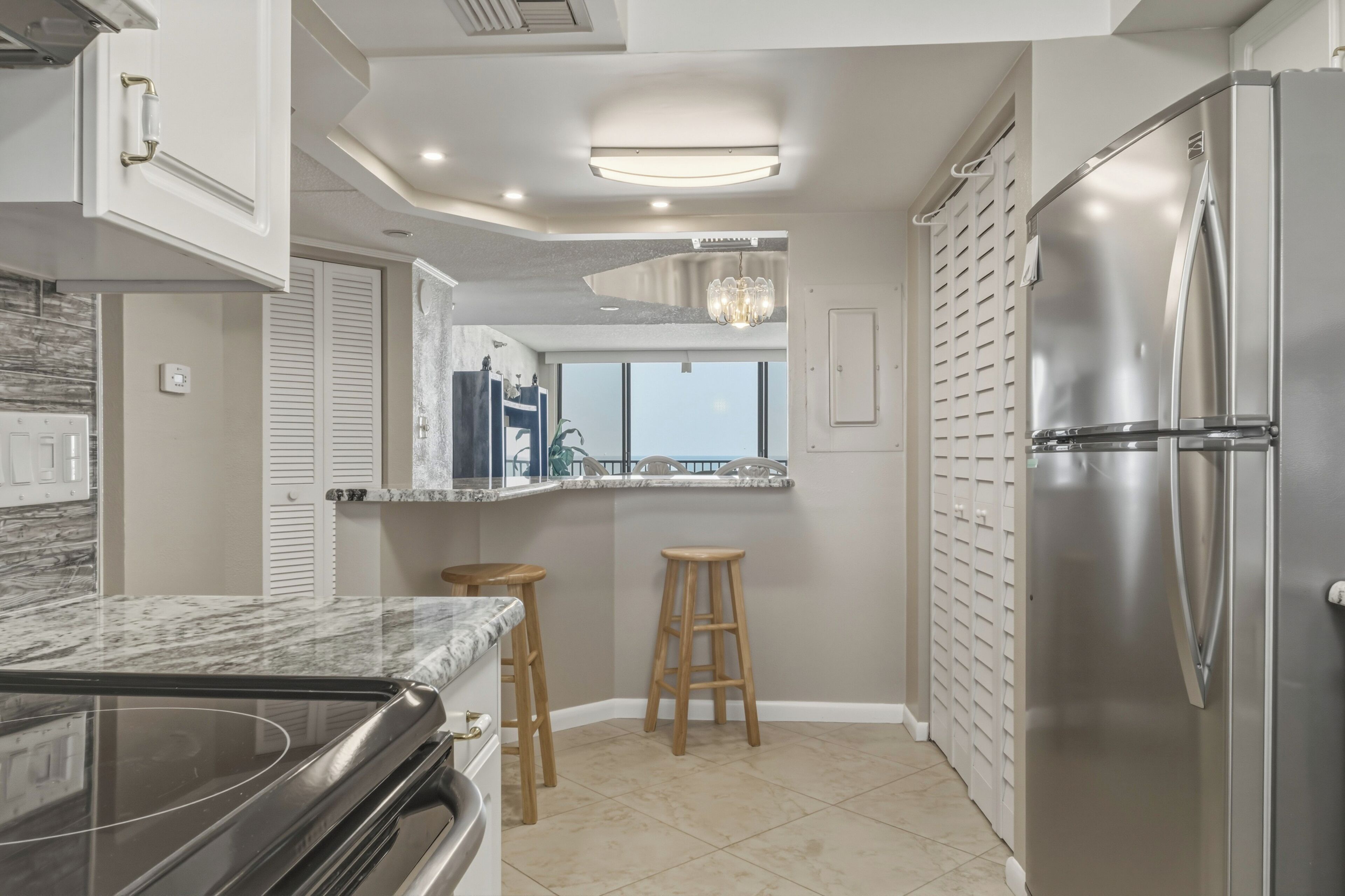 A kitchen with a stainless steel refrigerator, a built-in oven, and a marble countertop. There are two wooden stools and a window with a view.