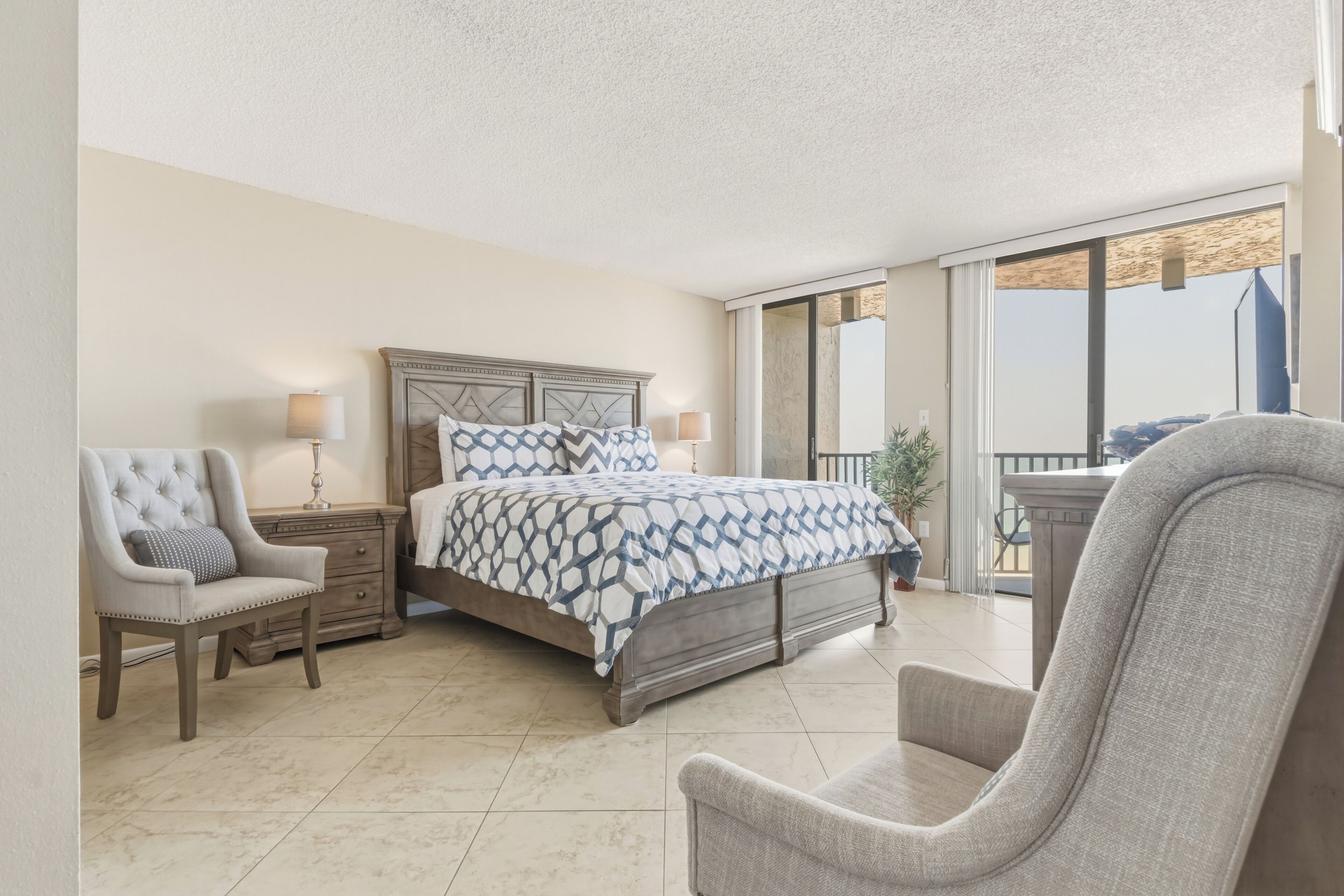 A bedroom with a bed, two bedside tables, a chair, and a balcony with a view.