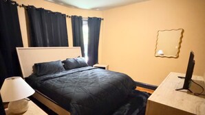 A bedroom with a bed, a bedside table with a lamp, a desk with a computer, and a window with curtains.