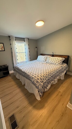 A bedroom with a bed, a window with curtains, a bedside table, and a headboard. - Cozy County Cottage- Pet friendly (Afton)