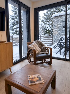 Ein gemütliches Zimmer mit einem Holzstuhl, einem Tisch mit einer Zeitschrift und großen Fenstern mit Blick auf eine verschneite Landschaft.