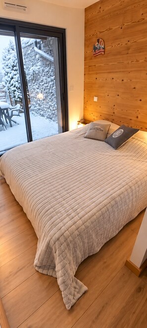 A bedroom with a large bed, a wooden headboard, and a view of a snowy outdoor area through a glass door. - travelski home premium - Résidence La Mariande - 3 Pièces 6 personnes N°0.4 (Les Deux Alpes)