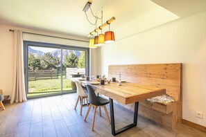 Uma sala de jantar moderna com mesa de madeira, cadeiras pretas e vista para uma paisagem montanhosa através de grandes portas de vidro.