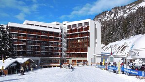 Ein Skigebiet mit einem großen Gebäude, mehreren Balkonen, einer Seilbahn und einem verschneiten Berg im Hintergrund.