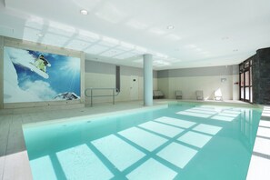 A swimming pool with clear water, surrounded by a tiled area with lounge chairs and a large mural of a snowy mountain. - travelski home select - Résidence Le Panoramic - 2 Pièces 4 personnes (Arâches-la-Frasse)