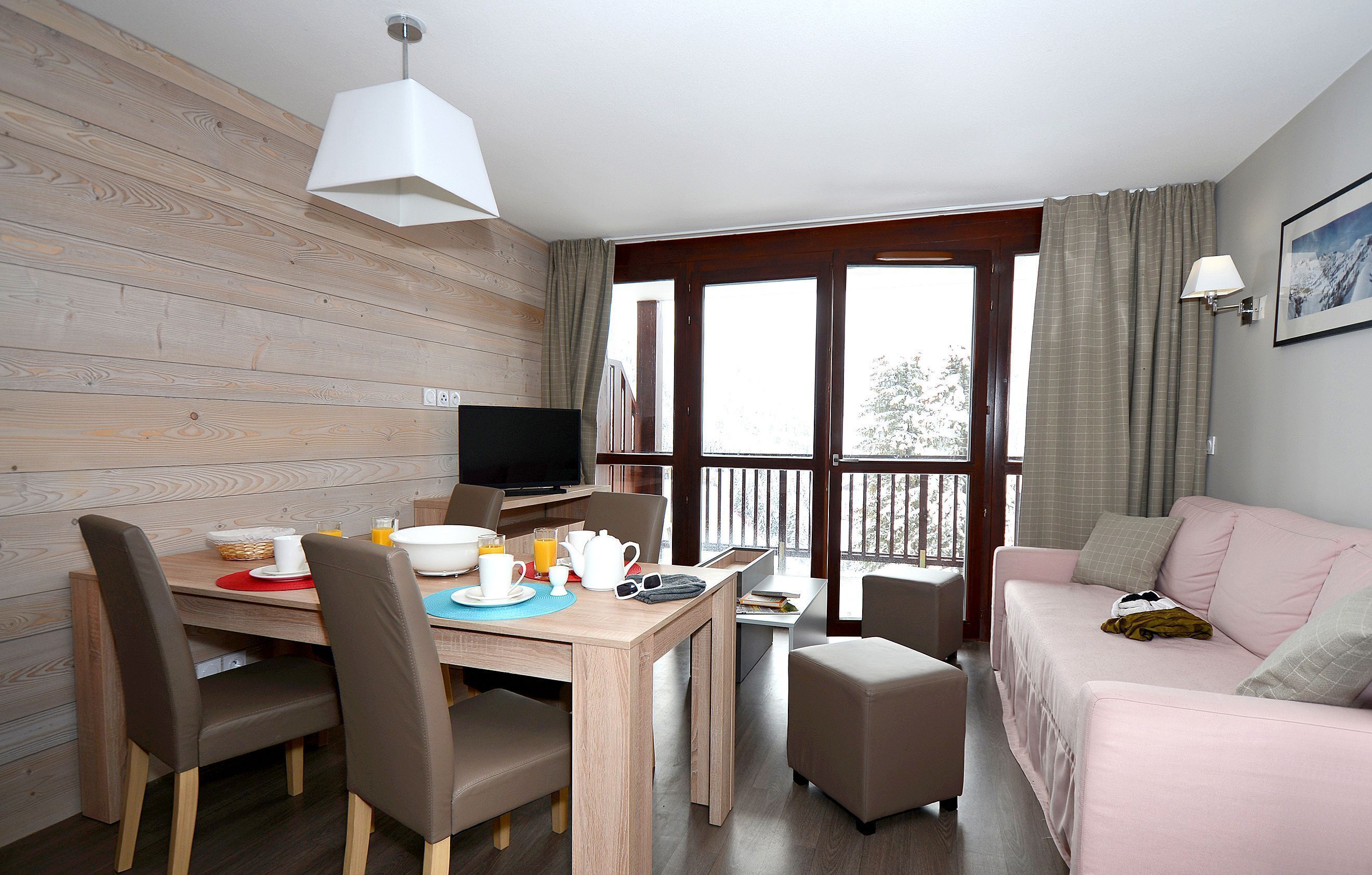 Une pièce comprenant une table à manger dressée pour le petit-déjeuner, un téléviseur, un canapé et un balcon avec des rideaux.
