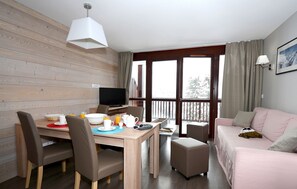 Et værelse med et spisebord dækket til morgenmad, et fjernsyn, en sofa og en balkon med gardiner.