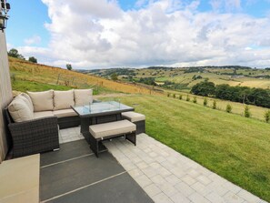 An outdoor patio with a wicker sofa and glass table, overlooking a hilly landscape. - The Stables (Halifax)