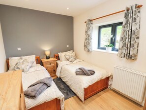 A bedroom with two beds, a bedside table with a lamp, a window with curtains, and a radiator. - The Stables (Halifax)