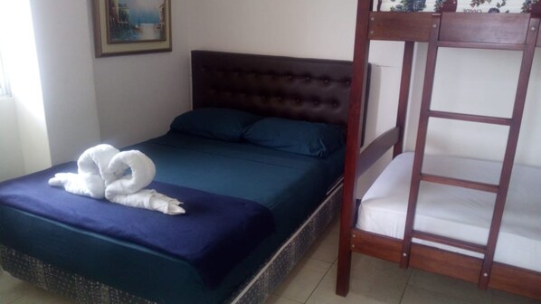 Una cama individual con cabecera oscura y colcha azul, una litera de madera a la derecha y un cuadro en la pared.