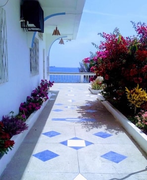 Un pasillo con suelo de baldosas, pared blanca con ventana y balcón con flores coloridas.