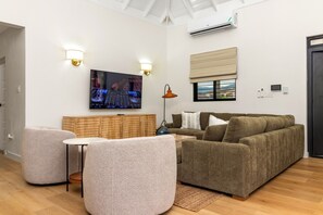 A modern living room with a flat-screen TV, a sofa, two armchairs, a wooden headboard, and a window with a view. - 5BR Luxe Retreat in Runaway Bay ~ Private Pool ~ Beach Access (Runaway Bay)