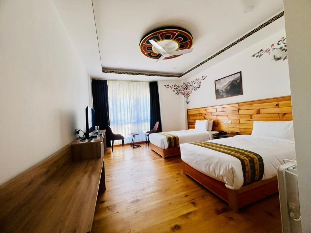 Quarto de hotel com duas camas, cabeceira de madeira, escrivaninha com cadeira, mesinha e janela com cortinas.