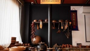 A traditional kitchen with a sign reading 'BHUTAN KITCHEN,' featuring wooden utensils, pots, and a large pot on a table. - Hotel River Valley Paro (Paro)