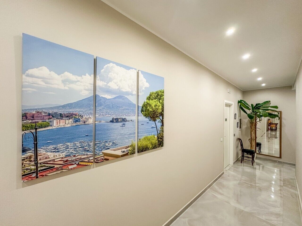 A hallway with a large painting of a coastal cityscape, a potted plant, and a mirror on the wall.