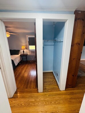A narrow hallway with a wooden floor leading to a bedroom with a bed and a desk with a lamp.