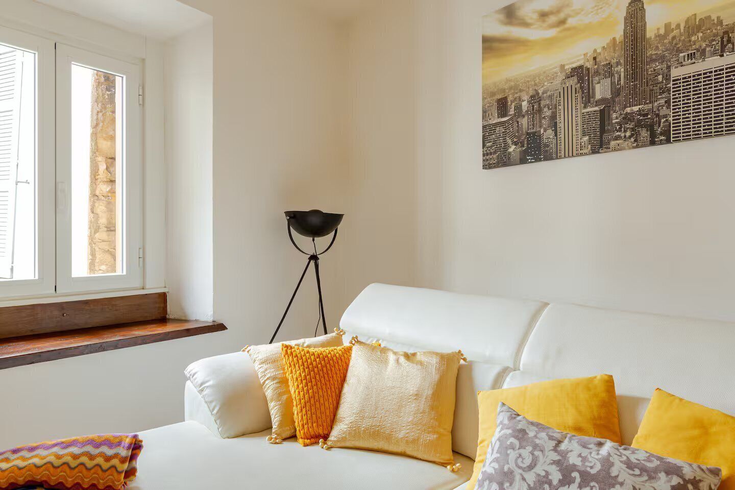 Un salon avec un canapé blanc, des coussins jaunes et blancs, un lampadaire et un tableau représentant un paysage urbain accroché au mur.