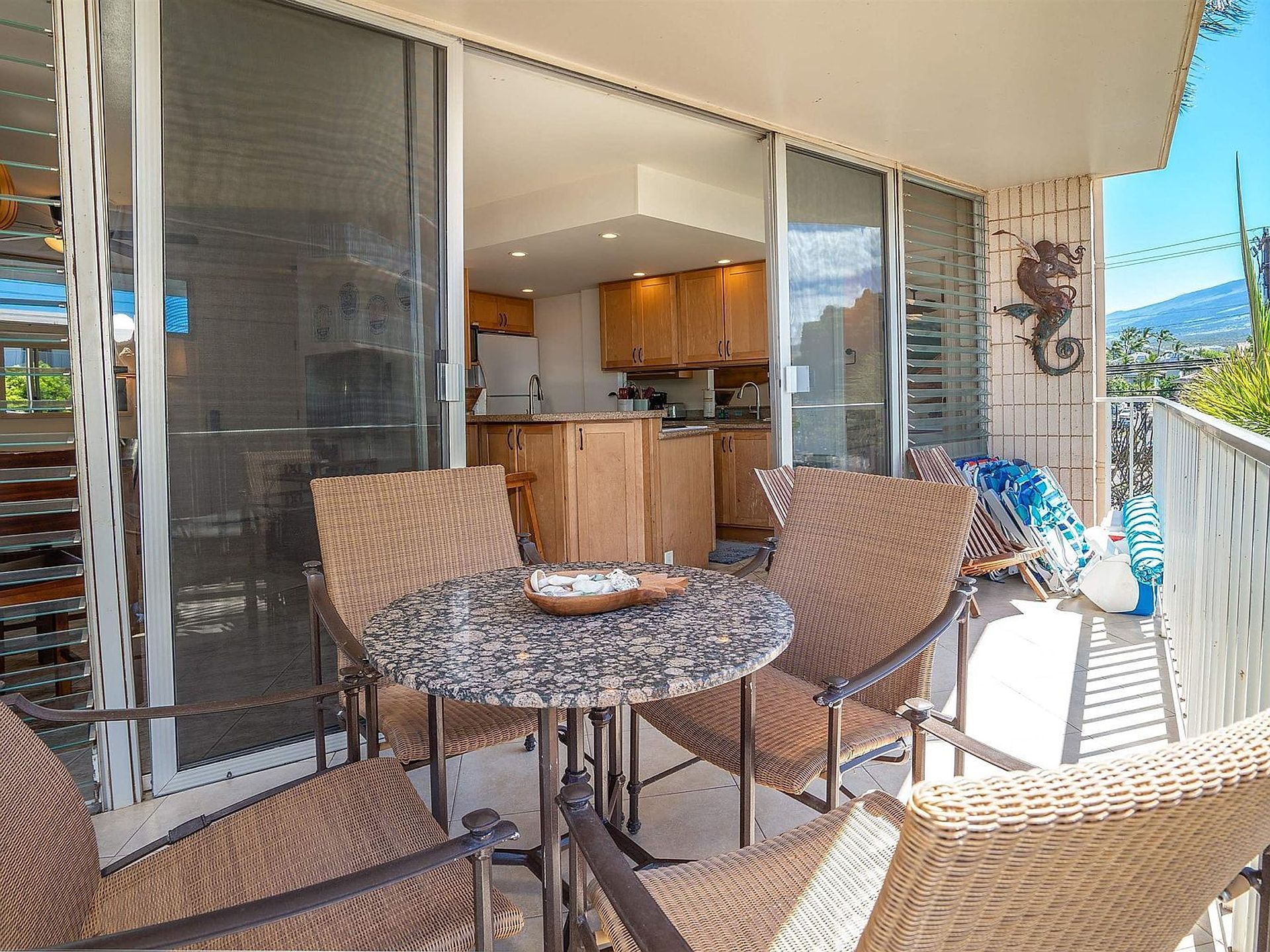 Een balkon met een ronde tafel en rieten stoelen, een schuifdeur naar een keuken en een decoratief kunstwerk van een zeepaard aan de muur.
