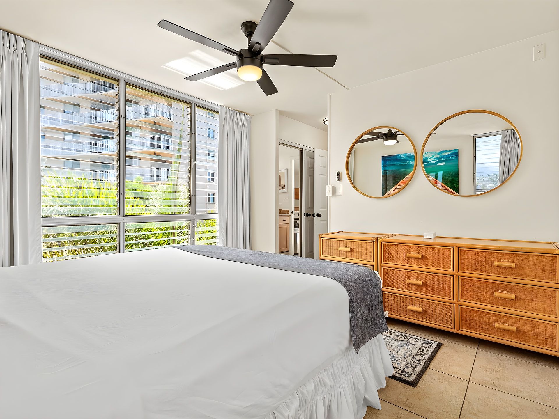 A bedroom with a bed, a ceiling fan, large windows, a wardrobe, and two wall mirrors.