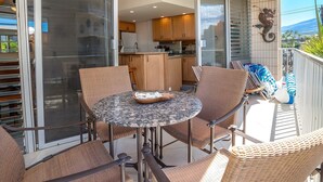 A balcony with a round table and wicker chairs, a sliding glass door leading to a kitchen, and a decorative wall piece. - Beachfront 4BR, 2 Condos for 12 in Kihei! (Kihei)
