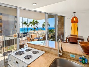 A kitchen with a view of the ocean, a dining area with a table and chairs, and a yellow pendant light. - Kihei, Kamaole Nalu, Spacious 4 BR, Beachfront, Sleeps 12 (Kihei)