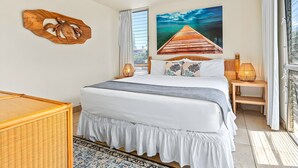 A bedroom with a bed, a ceiling fan, a wooden headboard, a wall-mounted artwork, a window with curtains, and a view of the ocean. - Beachfront 4BR, 2 Condos for 12 in Kihei! (Kihei)