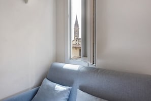 Eine Raumecke mit Fenster, einem grauen Sofa und Blick auf ein Gebäude mit einem Turm.