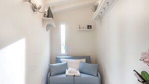 Um sofá-cama pequeno e azul claro com almofadas e uma toalha dobrada, uma porta branca e uma pequena prateleira com um porta-retrato e uma planta.