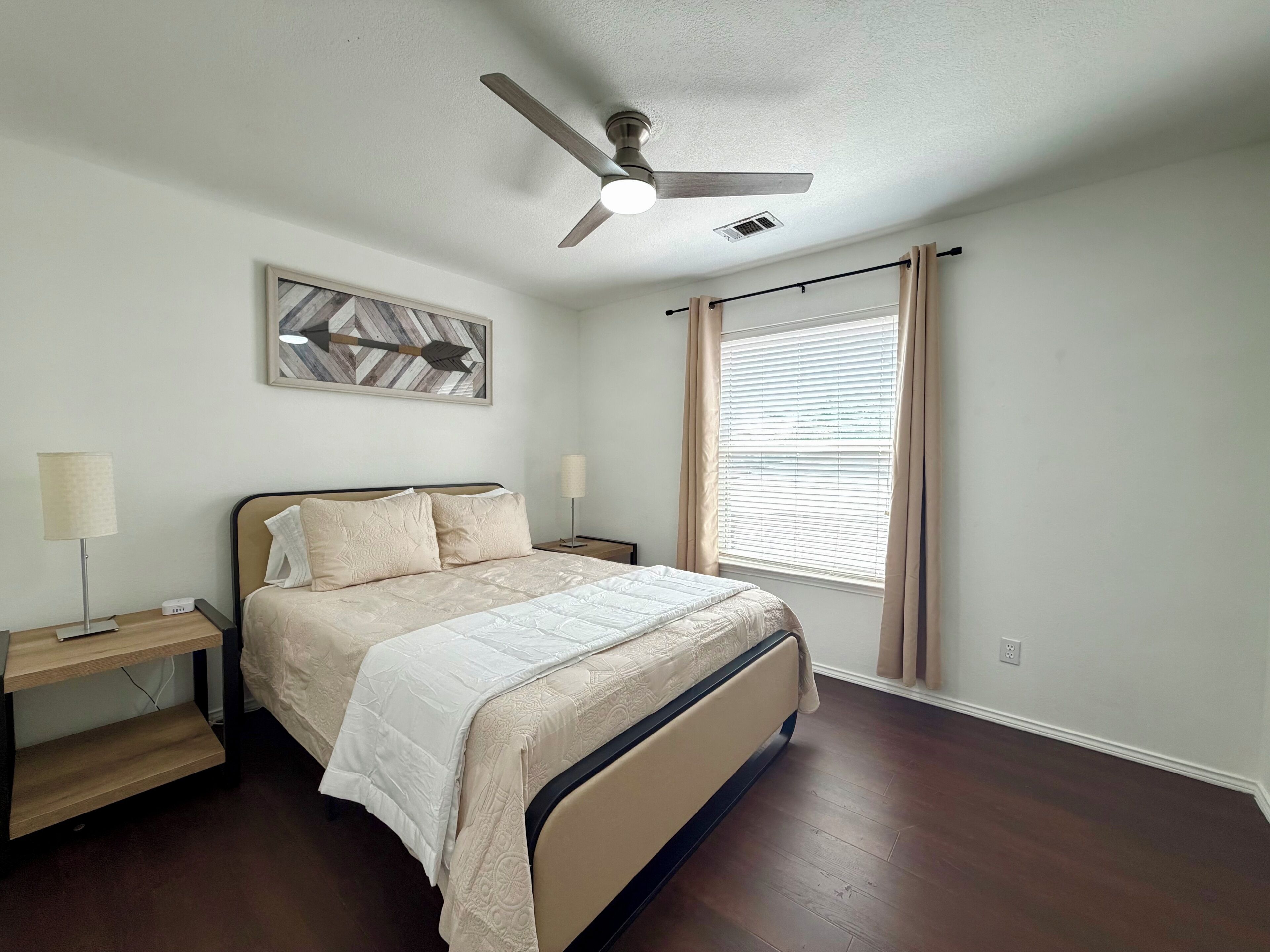 Quarto com cama, mesa de cabeceira, abajur, ventilador de teto, janela com cortinas e uma obra de arte na parede.