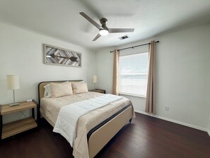A bedroom with a bed, a nightstand, a lamp, a ceiling fan, a window with curtains, and a wall-mounted artwork. - Luxe Family Retreat -Sleeps 10 - Large Home - Game Room -Private Back Yard (McKinney)