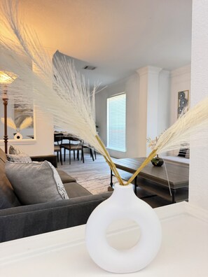 Ein Wohnzimmer mit einer weißen Vase auf einem Tisch, einem grauen Sofa und einem Essbereich im Hintergrund.