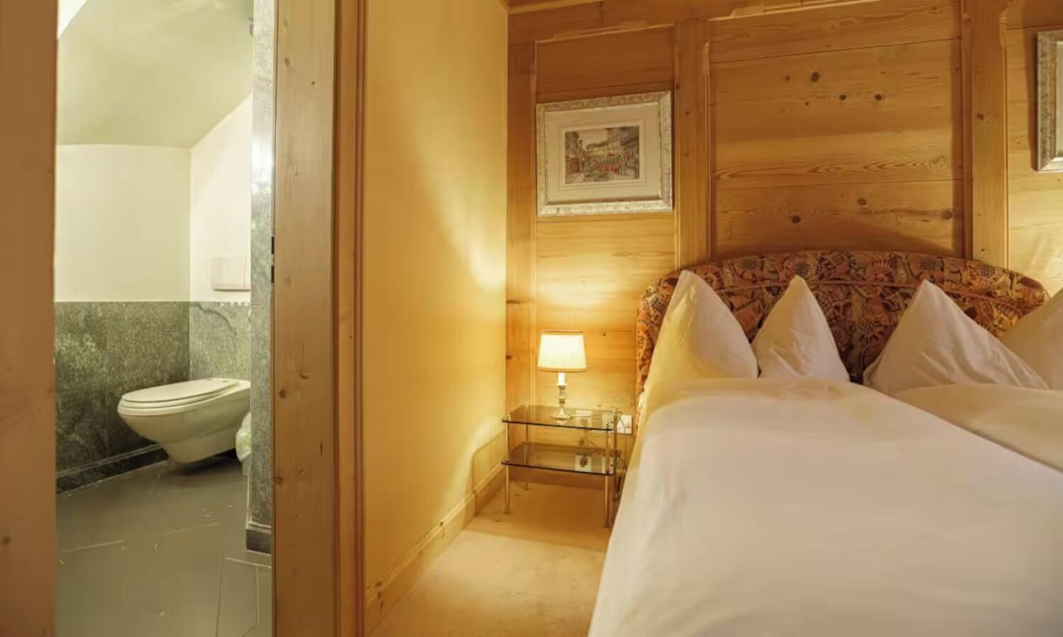 A wooden room with a bed, a bedside table with a lamp, and a framed picture on the wall.
