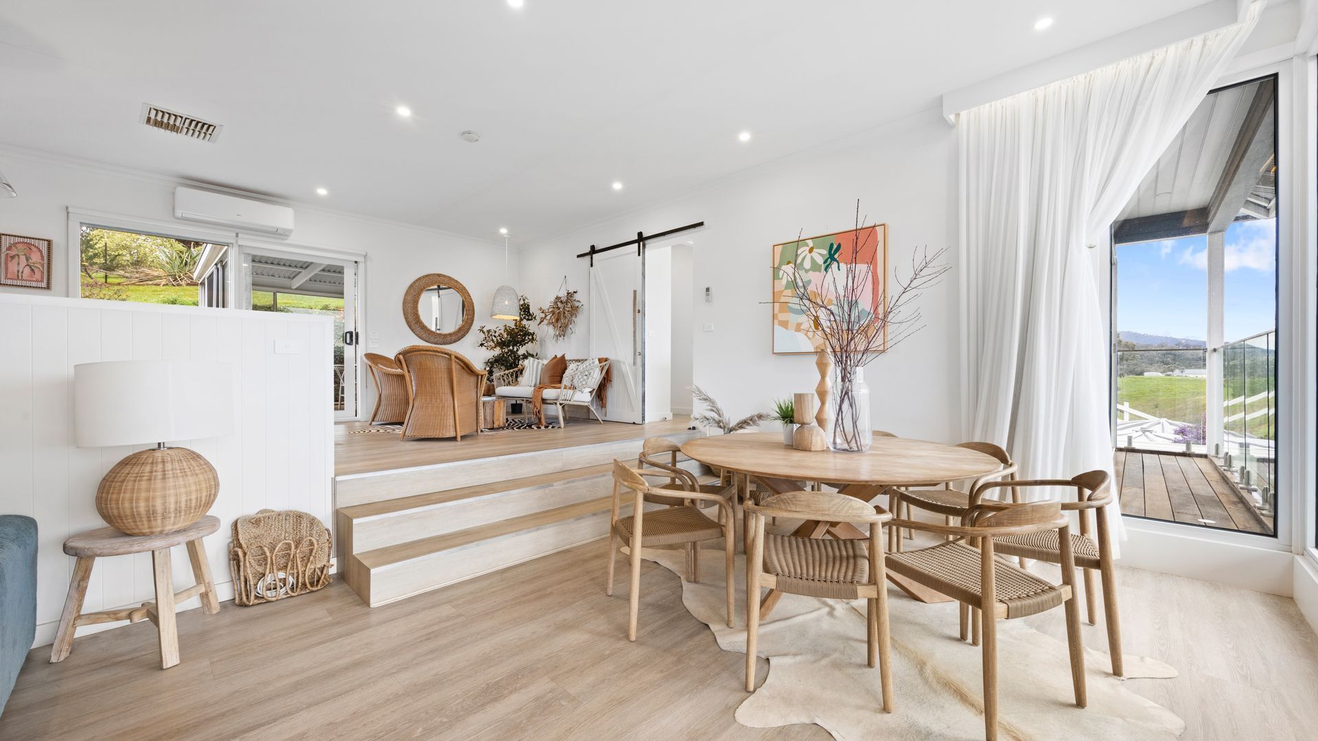 A modern interior with a dining area featuring a round table and chairs, a staircase leading to an upper level, and a large window offering a view of the outdoors.