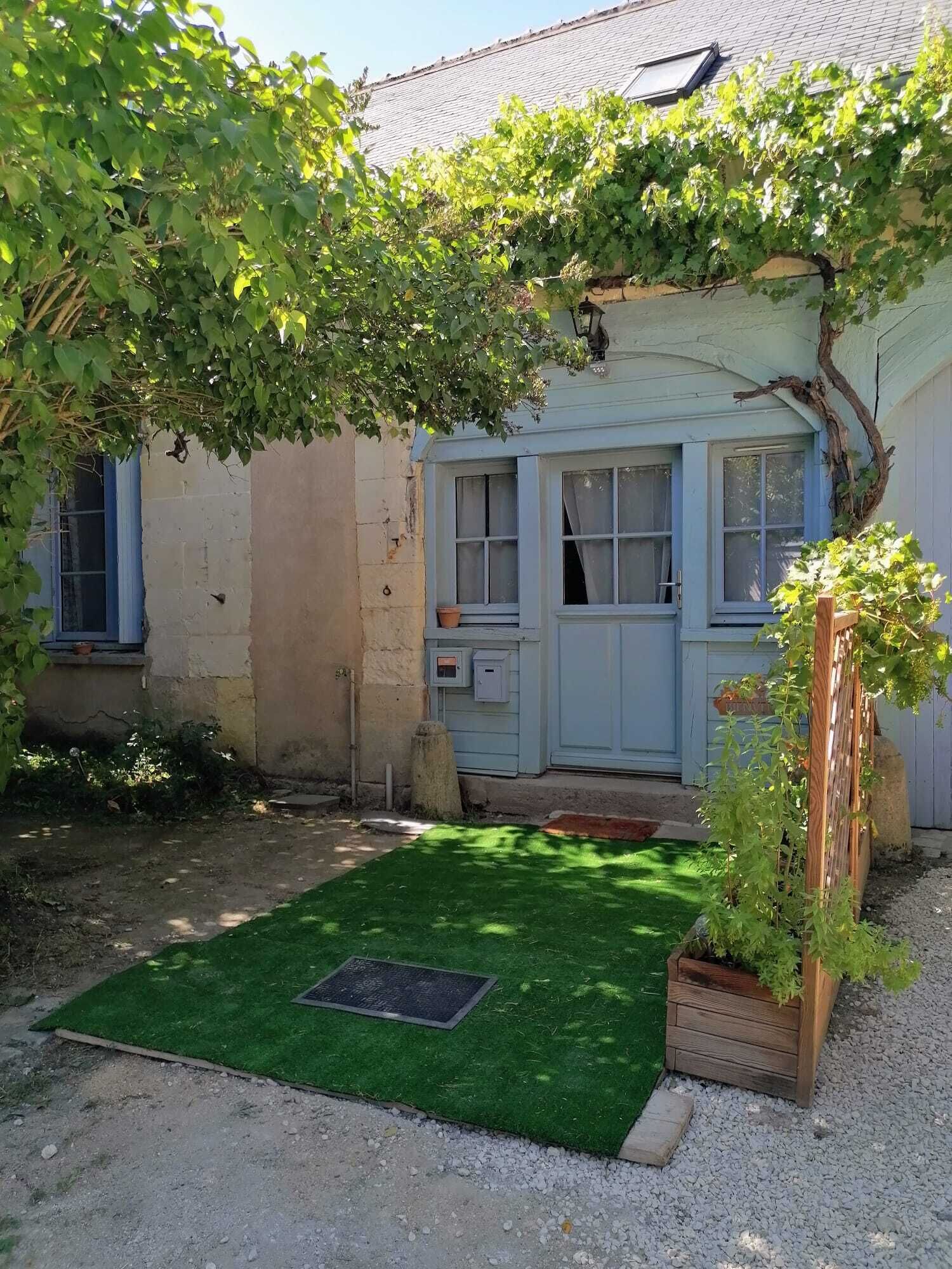 A small, single-story building with a blue door and windows, surrounded by greenery and a small lawn.
