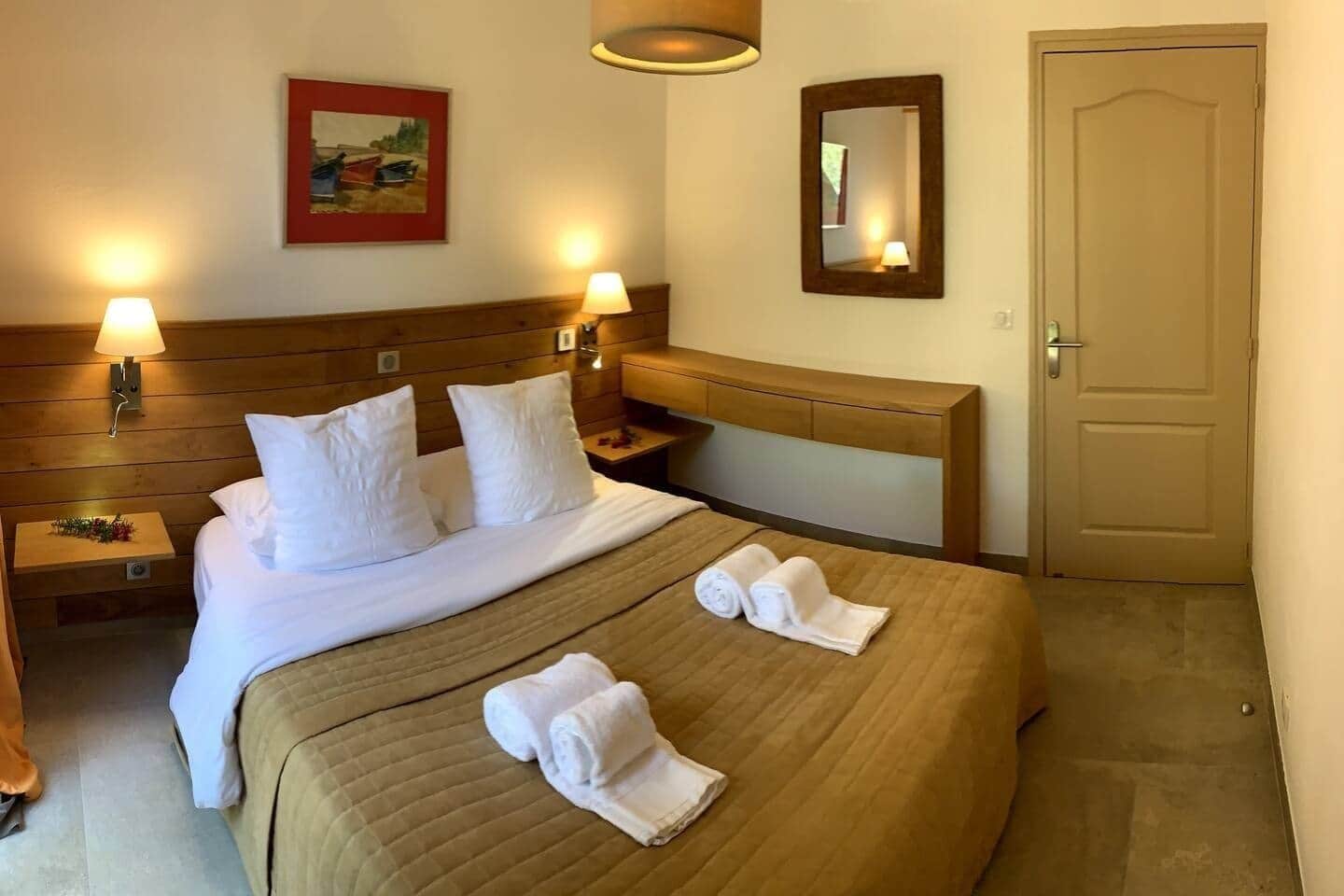 Habitació d'hotel amb un llit, dues làmpades de nit, una taula de nit, un capçal de llit de fusta, una porta i un mirall fixat a la paret.