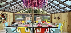 A covered outdoor dining area with a wooden structure, two hanging chairs, and a table set for two with colorful chairs and plates.
