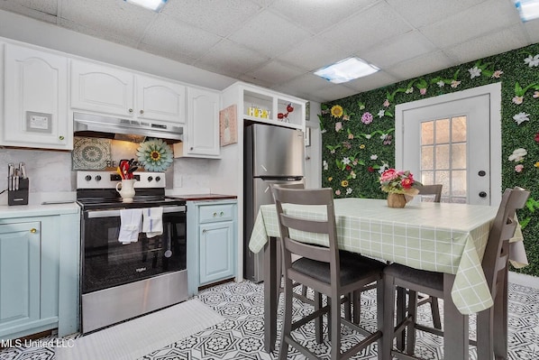 A kitchen with white cabinets, a stainless steel stove, and a refrigerator. There is a dining table with chairs and a flower vase.