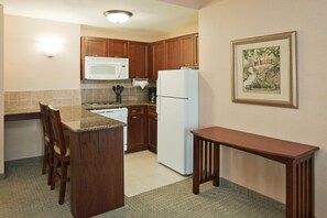 A compact kitchen with wooden cabinets, a white refrigerator, a microwave, and a coffee maker. There is a small island with a chair, and a framed picture on the wall.