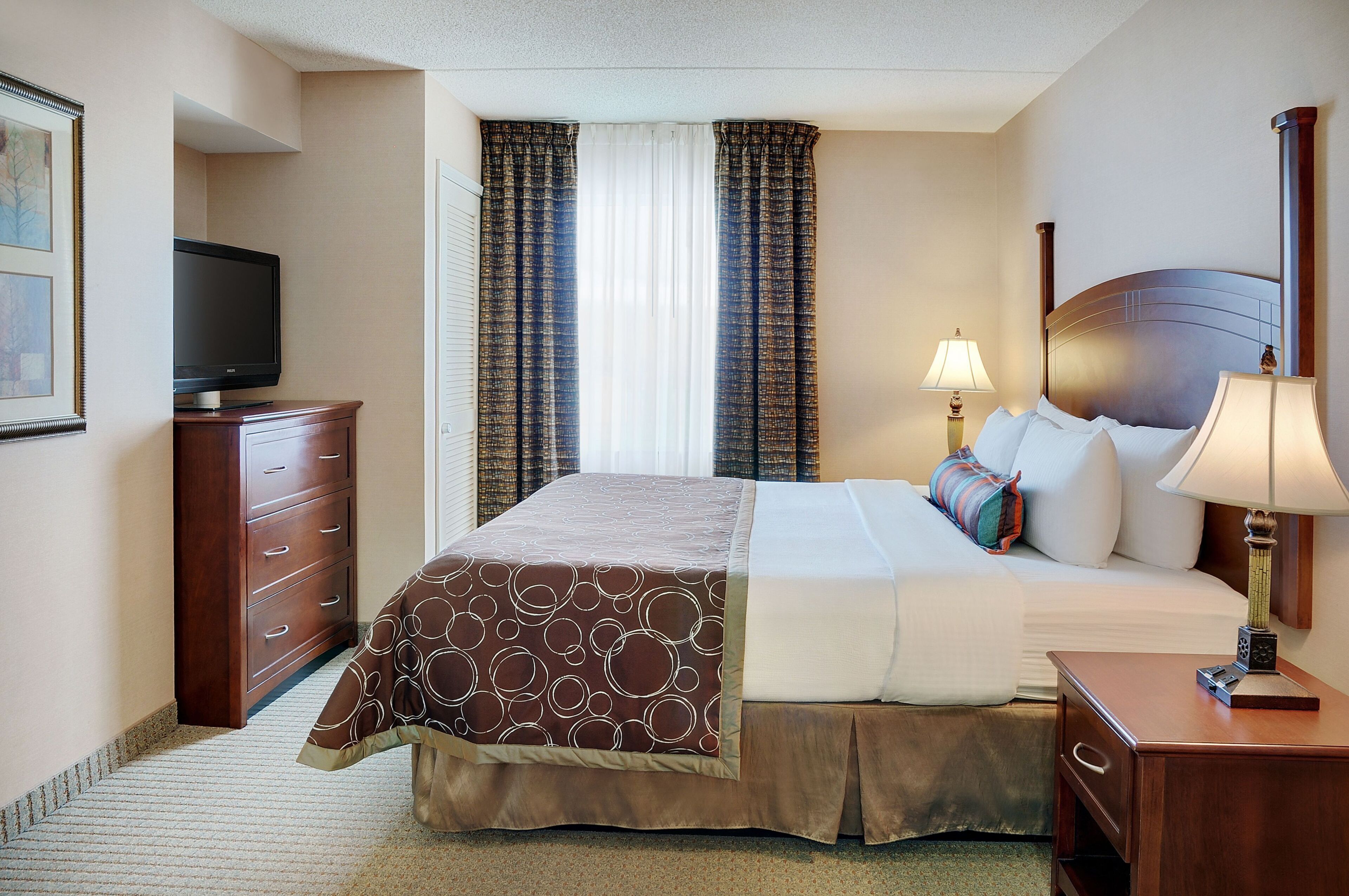 A hotel room with a bed, a television, a desk with a lamp, and a window with curtains.
