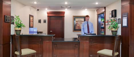 A reception area with a wooden reception desk, two chairs, a man standing behind the desk, and a door with a framed picture above it.