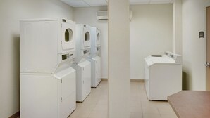 A laundry room with multiple white washing machines and dryers, a tiled floor, and a plain wall with an air conditioning unit.