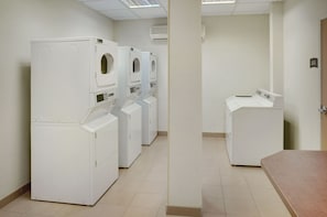 A laundry room with multiple white front-loading washers and dryers, a tiled floor, and a plain wall with an air conditioning unit.