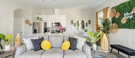 A modern living room with a grey sofa, a kitchen in the background, and a wall decorated with greenery and woven baskets.