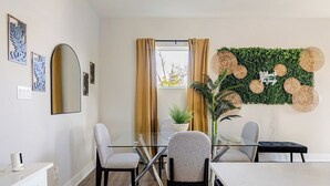 A modern dining area with a glass table, two chairs, a potted plant, and a wall-mounted decorative panel. - 3BR Gated Community Houston Home  + Parking (Houston)